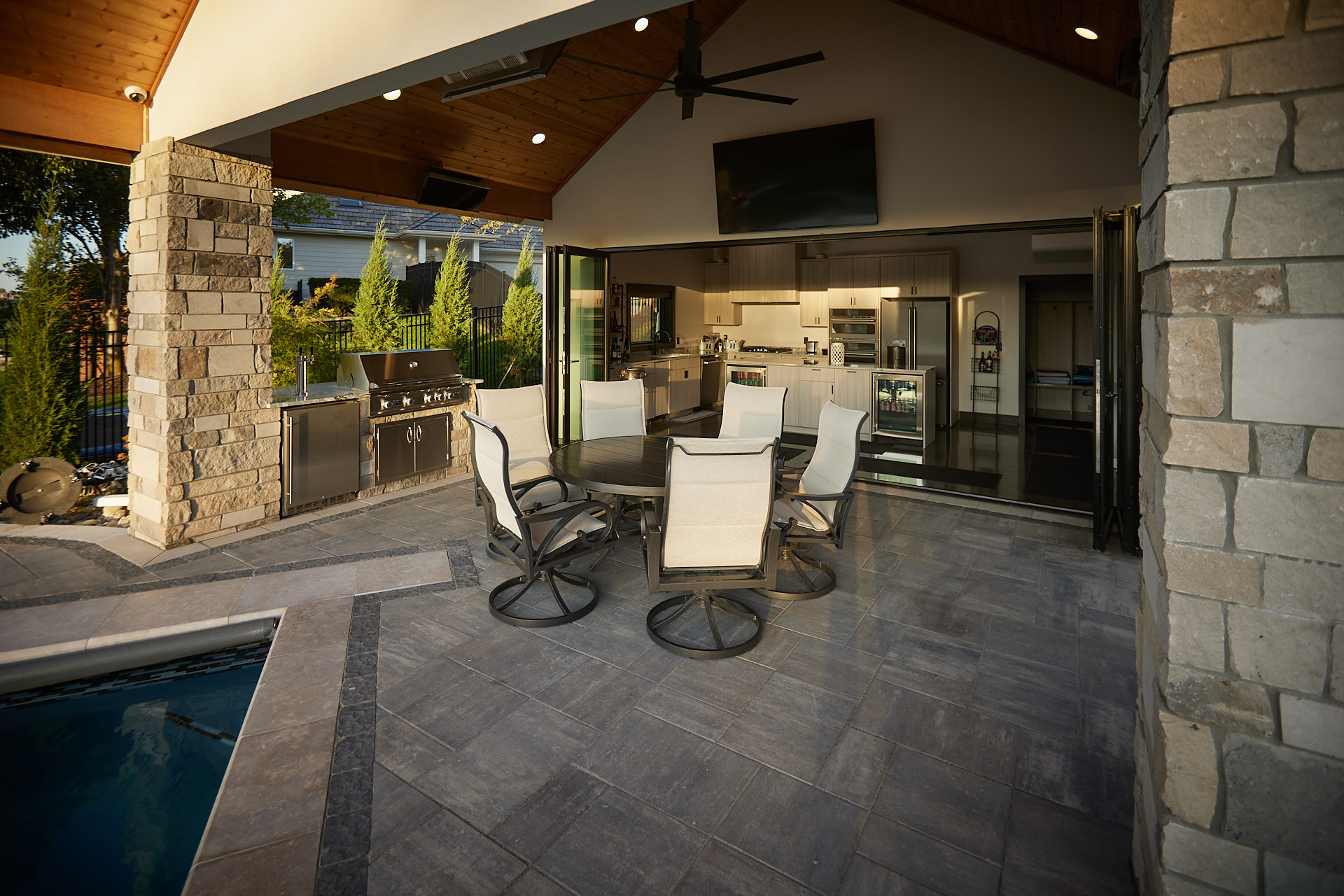 outdoor patio designed by indigenous landscaping of omaha ne. the patio has chairs and a table under a covered patio with a tv and outdoor kitchen